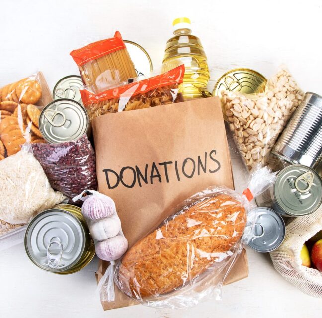 A paper grocery bag marked Donations surrounded by canned goods, pasta, cereal and bread